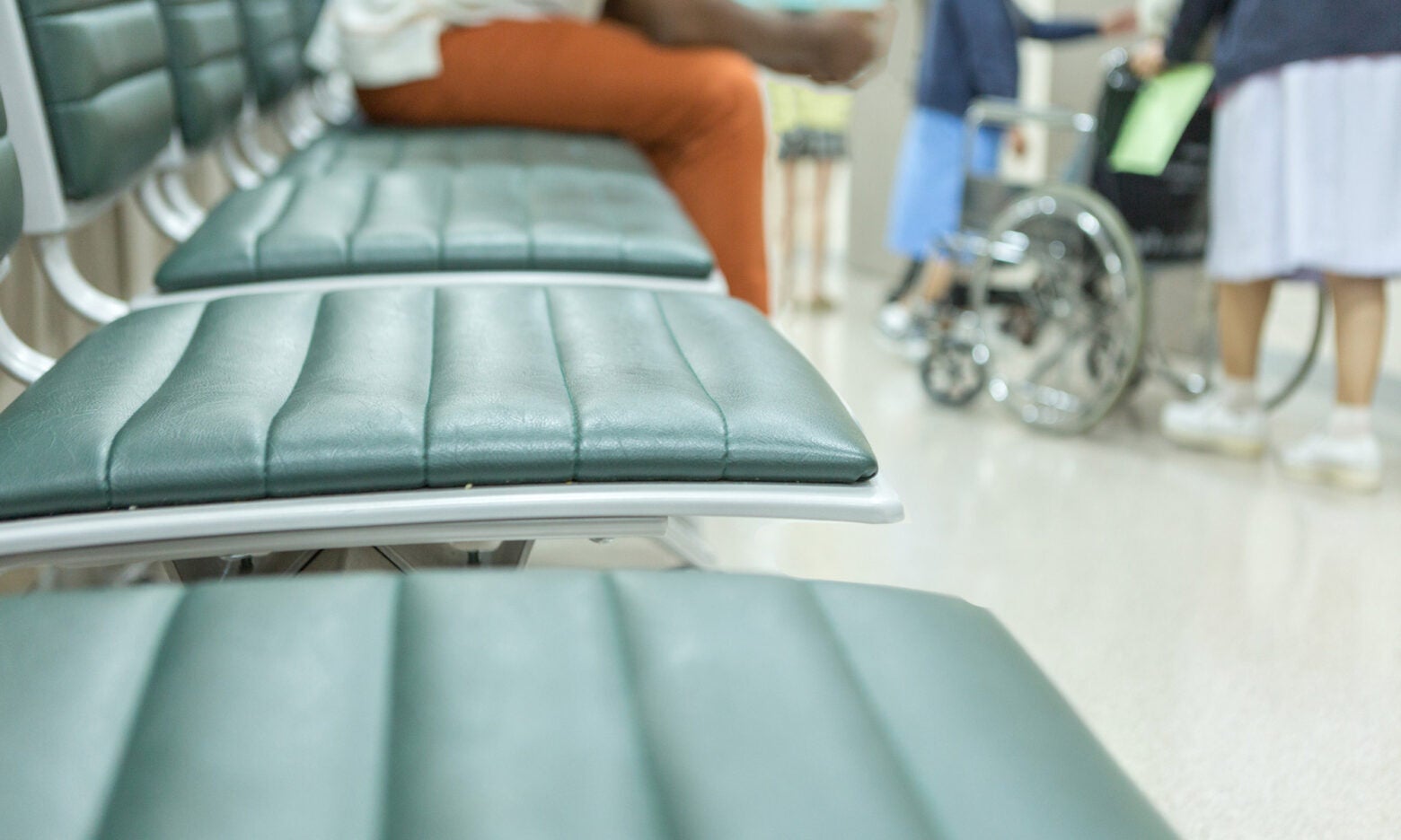 A man sits in a row of waiting room seats while nurses and wheel chairs move through a busy hallway at a hospital.