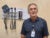 William Roberson stands in a medical office and looks at the camera. He wears a blue polo shirt and has a name tag around his neck. Behind him are wall-installed standard medical devices.